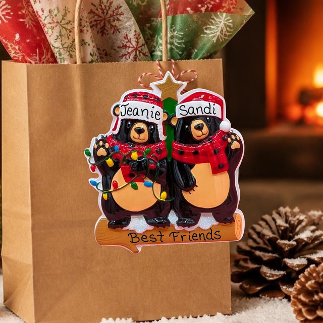 A personalized Christmas ornament shaped like two bear best friends hanging from a kraft gift bag. The ornament features two black bears wearing red Santa hats and red plaid scarves, with one bear holding a string of multicolor Christmas lights and the other holding a green tree branch.