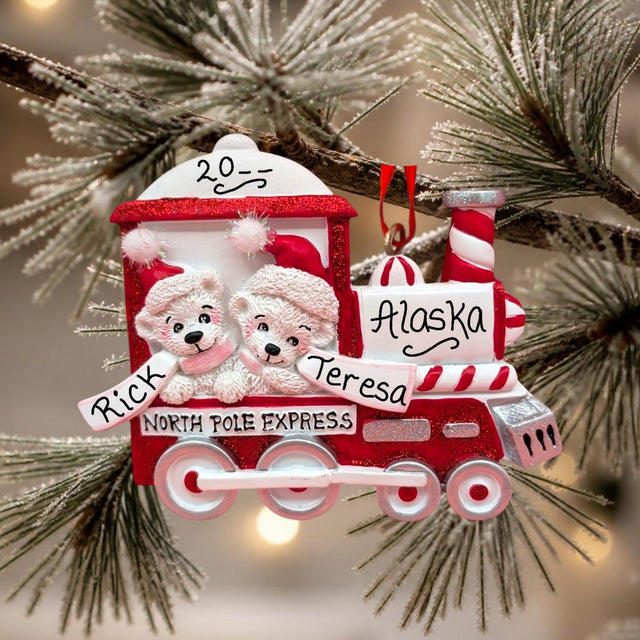 A personalized Christmas train ornament hanging on a snowy pine branch. The red and white ornament features two polar bear children in Santa hats, the words 'North Pole Express,' 'Alaska,' and the names 'Rick' and 'Teresa,' with the number '2015' on the roof.